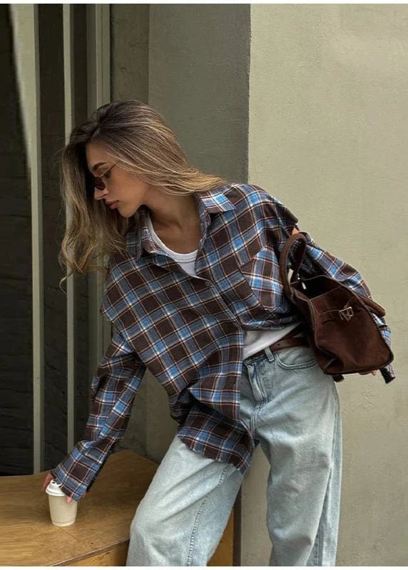 Woman in plaid shirt and jeans with a brown bag, standing by a wooden table.