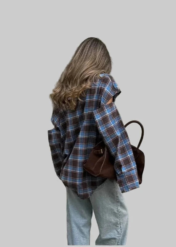 Person wearing a plaid shirt and holding a brown bag against a neutral wall.