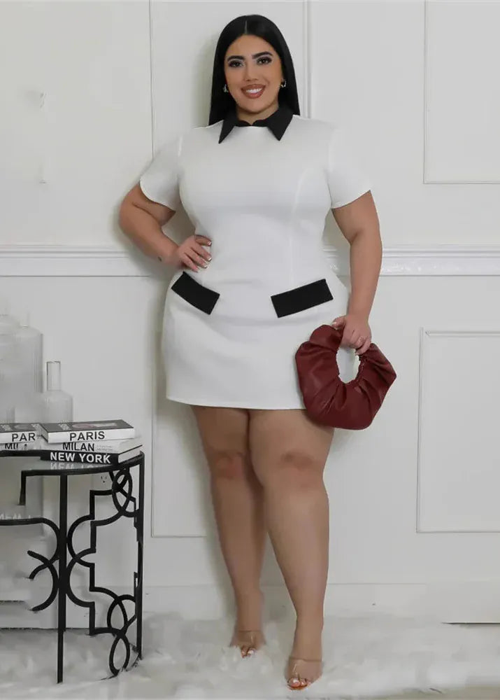 Woman in a white dress with black belts holding a red clutch against a white wall.