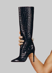 Black crocodile-patterned knee-high boot held by a hand against a red background