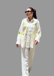 Woman wearing a white shirt with lemon pattern and white pants against a wooden wall.