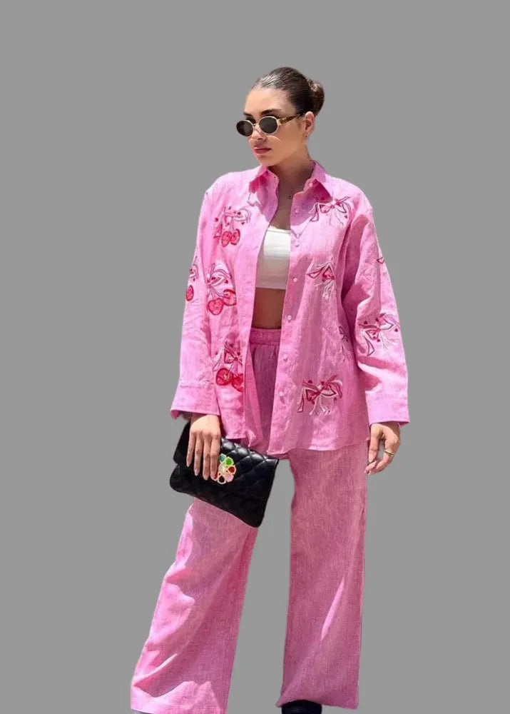 Woman in a pink outfit with embroidery standing against a wooden wall.