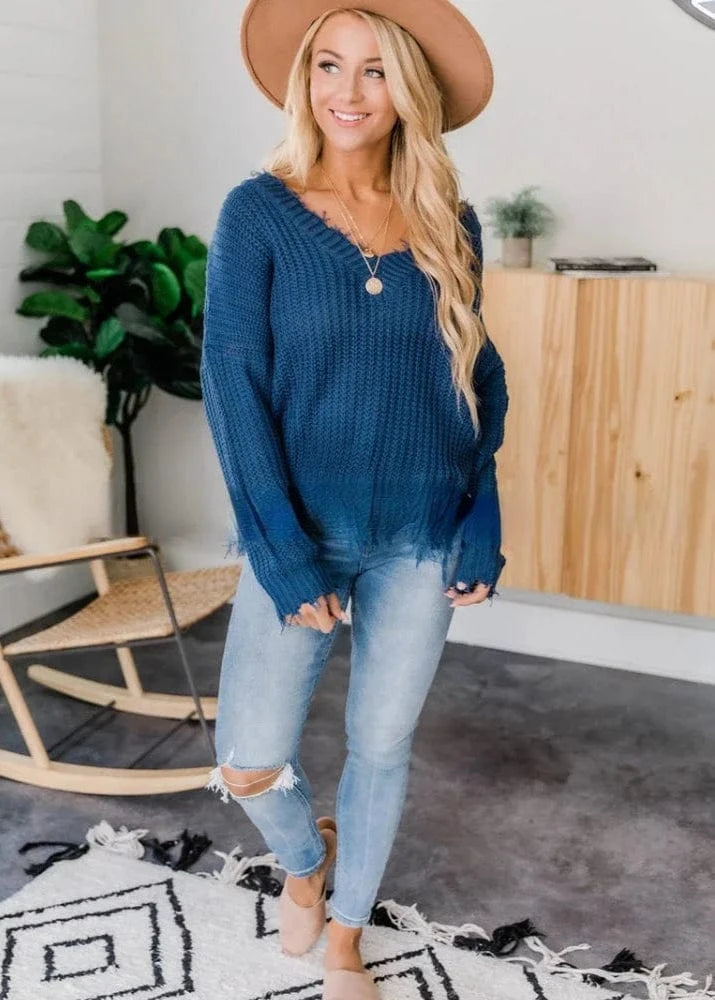 Woman wearing a blue sweater and light blue jeans in a room with a plant and wooden cabinet.