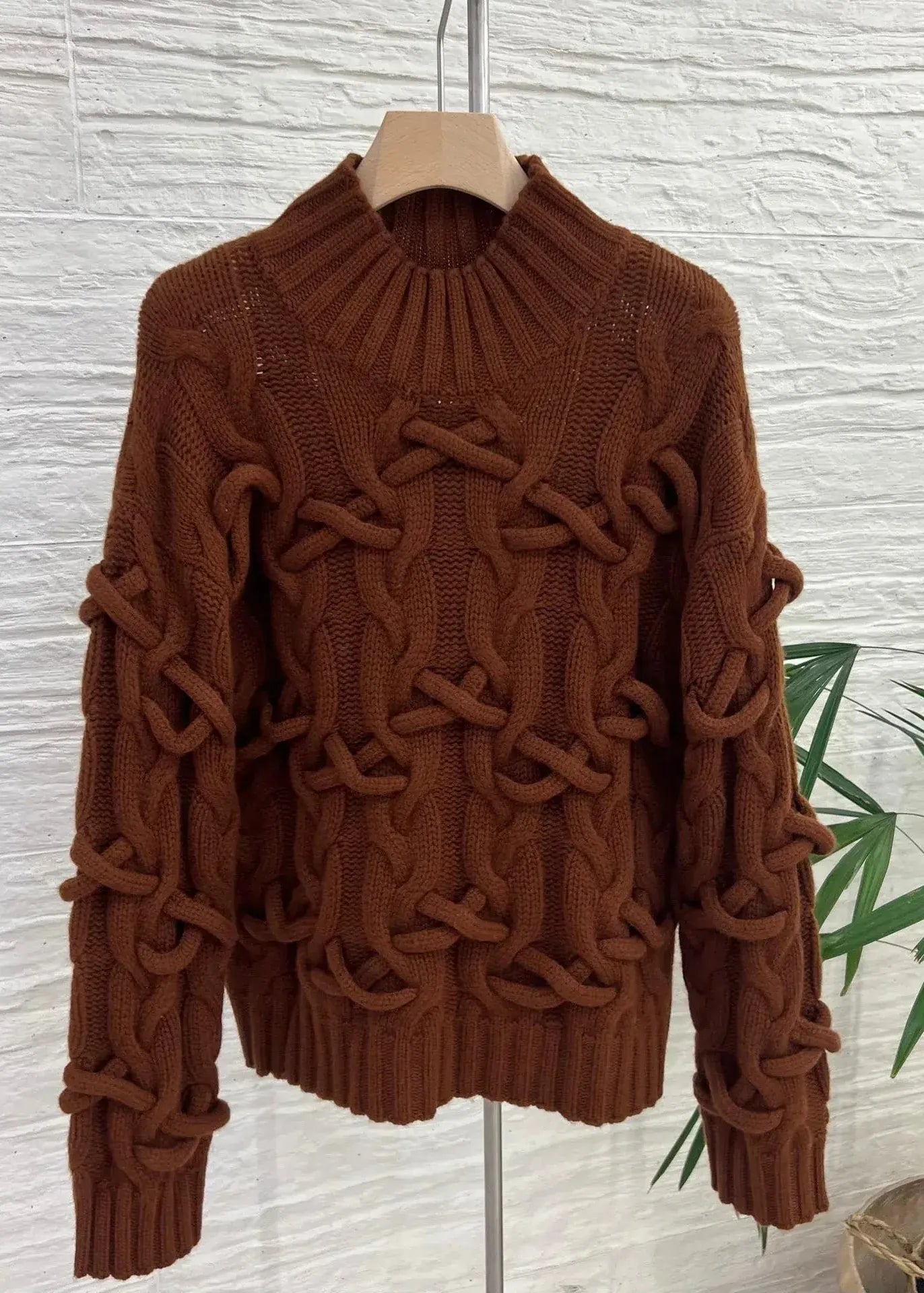 Brown cable knit sweater on a hanger against a textured white wall.