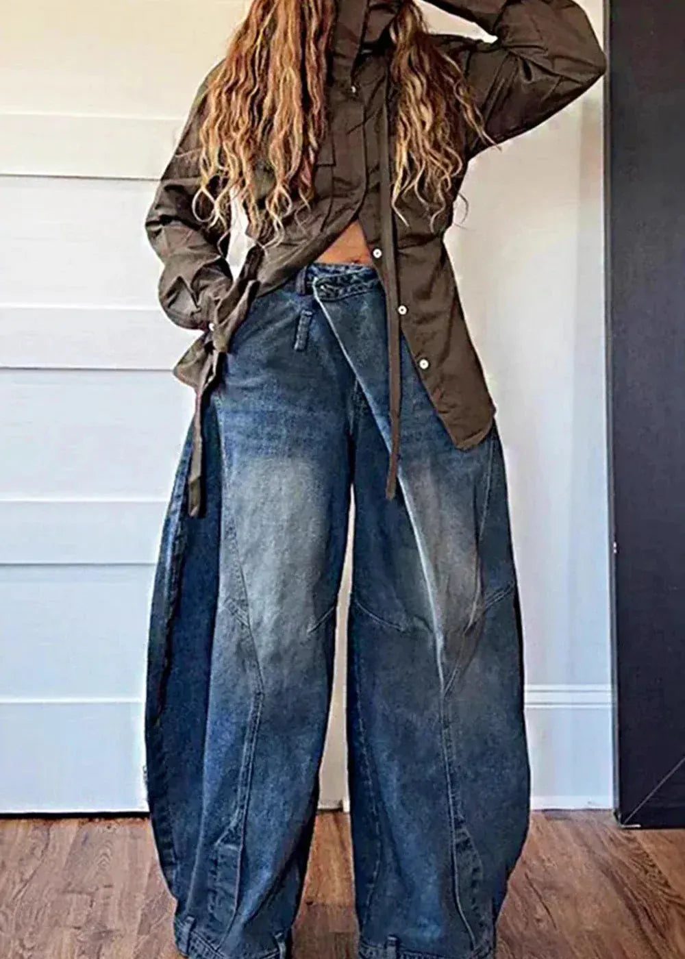 Person wearing a green jacket and blue jeans standing against a white wall.