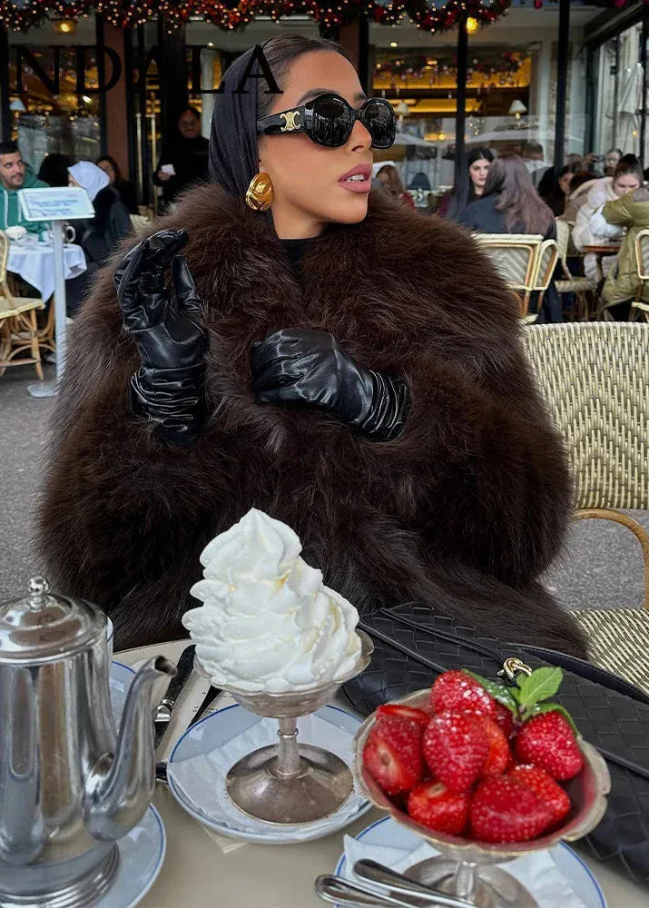 Woman in a fur coat and sunglasses sitting at an outdoor cafe table with a dessert and strawberries.