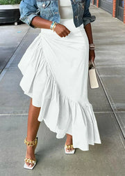 A woman wearing a white ruffled asymmetrical midi skirt with a denim jacket, white top, and gold jewelry, standing on a concrete sidewalk.