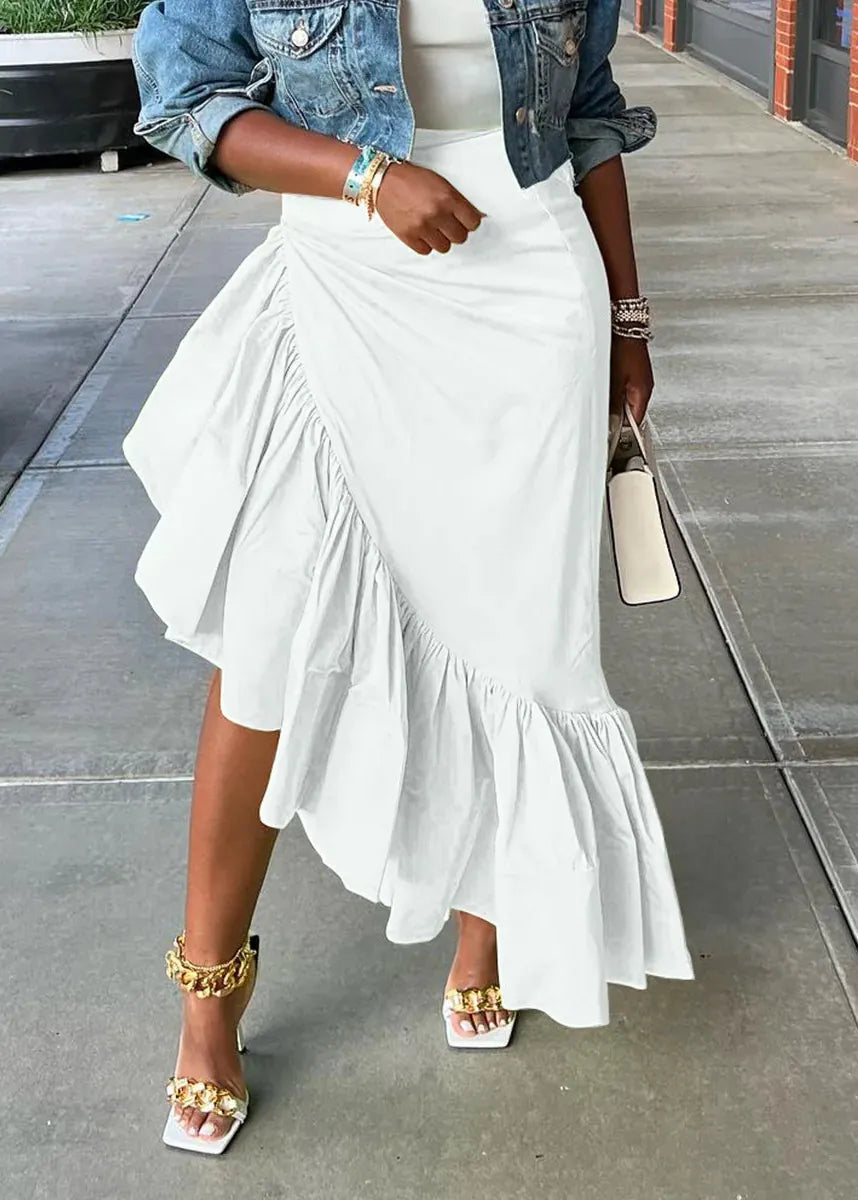 A woman wearing a white ruffled asymmetrical midi skirt with a denim jacket, white top, and gold jewelry, standing on a concrete sidewalk.