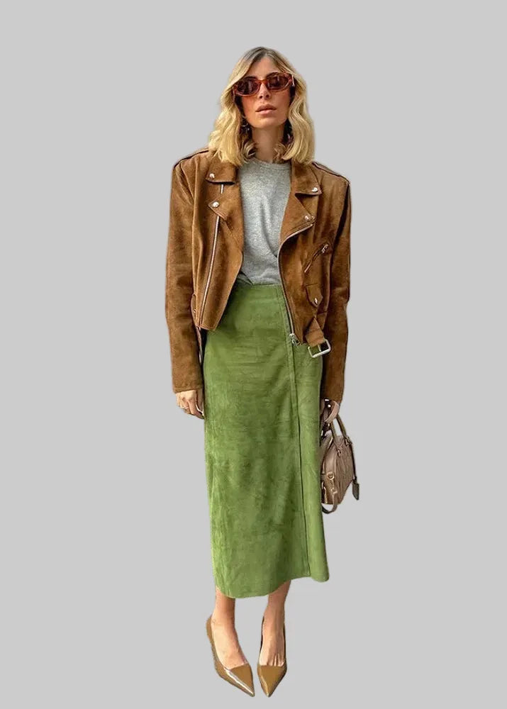 A woman stands outdoors in front of a building with arches, wearing a brown suede moto jacket over a gray top and an olive green midi skirt, with tan pointed-toe heels and a small taupe handbag.