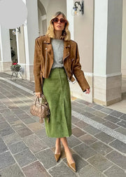 A woman stands on a stone walkway in front of a building with white columns, wearing a brown suede moto jacket over a gray top and an olive green midi skirt. She is holding a beige woven handbag and wearing pointed-toe heels.