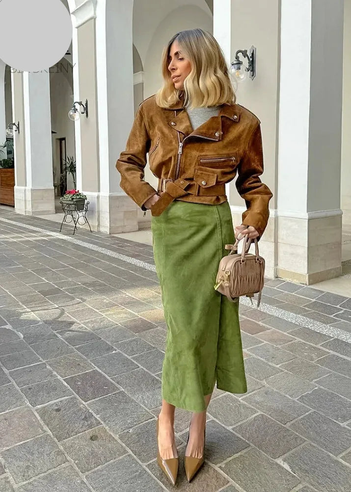 A woman stands in a colonnade wearing a brown suede moto jacket and an olive green midi skirt, holding a small brown handbag. The image has a watermark logo 'SR GINGRON' in the top left corner.