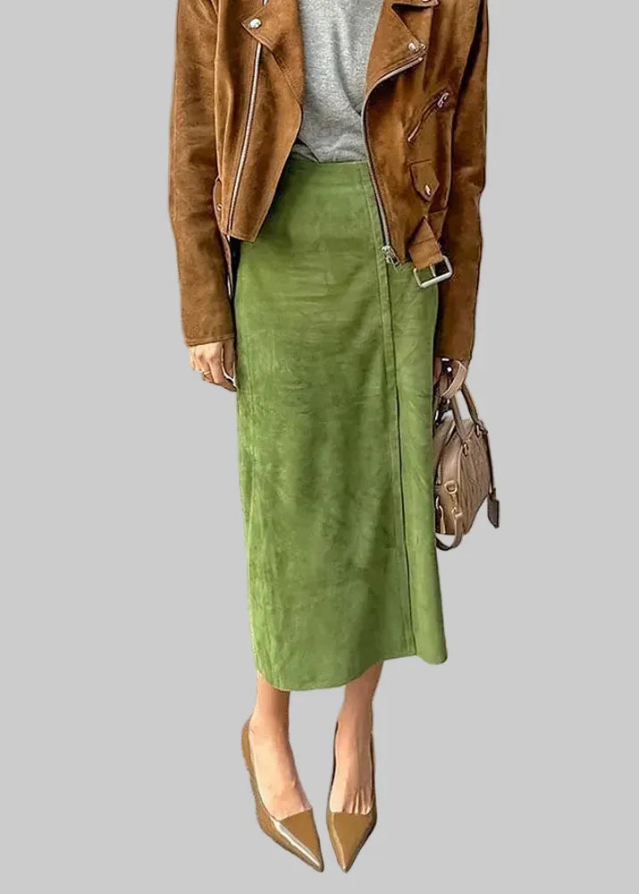 A woman wearing a brown suede moto jacket, a gray knit top, and a green midi skirt, standing on a stone paved street. She is holding a brown handbag and wearing pointed-toe heels.