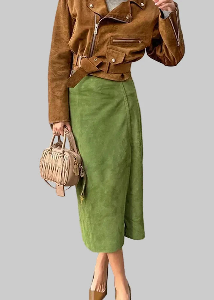 A woman wearing a brown suede moto jacket and an olive green midi skirt, holding a beige fringed handbag on a stone-paved outdoor walkway. The brand logo 'SR SINGREINY' is visible in the top left corner.