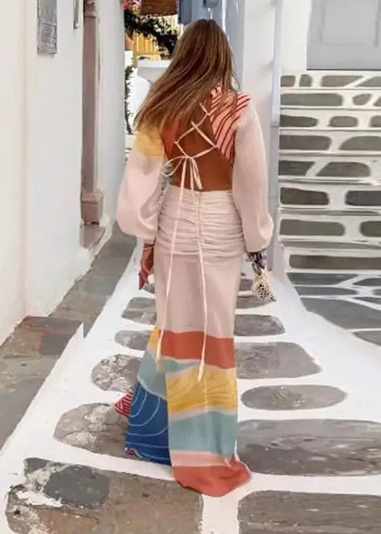 A woman viewed from behind wearing a long, multicolored bohemian maxi dress with a brown wrap-style back and layered sheer sleeves, walking down a white-walled alleyway with stone steps.