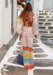 A woman viewed from behind wearing a long, multicolored bohemian maxi dress with a brown wrap-style back and layered sheer sleeves, walking down a white-walled alleyway with stone steps.