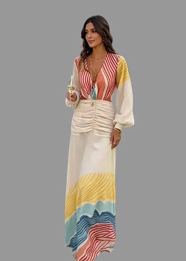 A woman stands outdoors on a grassy lawn wearing a long, multicolored maxi dress with a striped bodice and a wavy, layered skirt. She is holding a glass of white wine.
