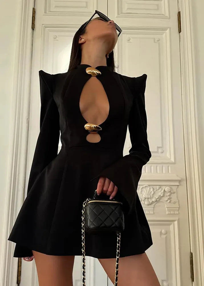 Person wearing a black dress with cut-out design, holding a black handbag against a white wall.