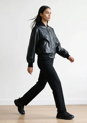 A woman walking in profile wearing a black leather bomber jacket, black pants, and black sneakers against a plain white wall and light wood floor.