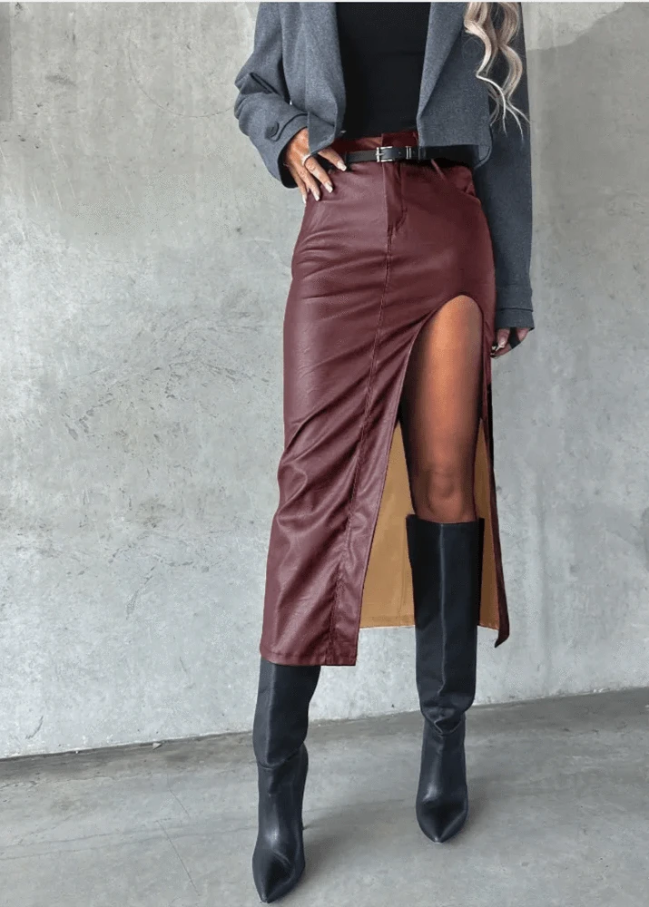 Person wearing a burgundy leather skirt with a high slit and black knee-high boots against a gray concrete wall.
