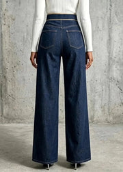 A model shown from the back wearing dark blue wide-leg denim jeans and a white ribbed long-sleeve top, standing against a textured concrete wall.