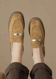 A pair of tan suede loafers with a small gold metal bar detail across the top, worn with dark brown trousers. The photo is taken from a top-down perspective showing the feet and legs against a solid beige background.