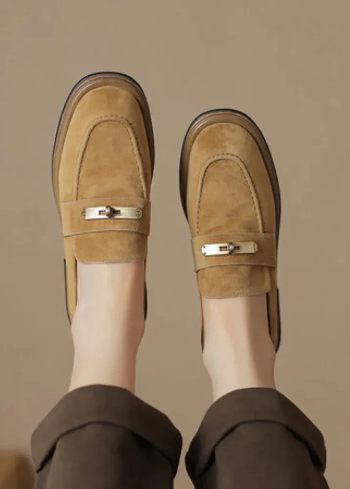 A pair of tan suede loafers with a small gold metal bar detail across the top, worn with dark brown trousers. The photo is taken from a top-down perspective showing the feet and legs against a solid beige background.