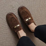 A pair of women's brown suede loafers with a gold-tone metal accent on the strap, shown being worn with black pants on a textured beige carpet.