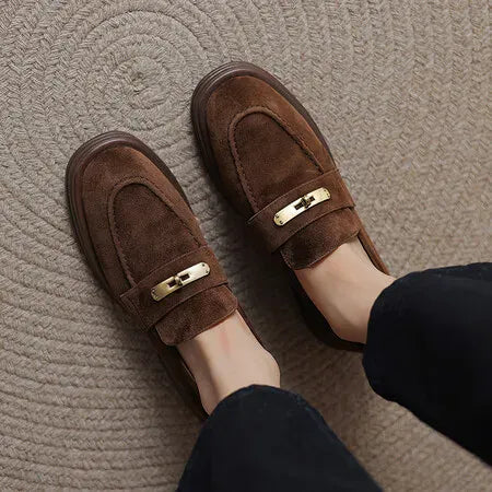 A pair of women's brown suede loafers with a gold-tone metal accent on the strap, shown being worn with black pants on a textured beige carpet.