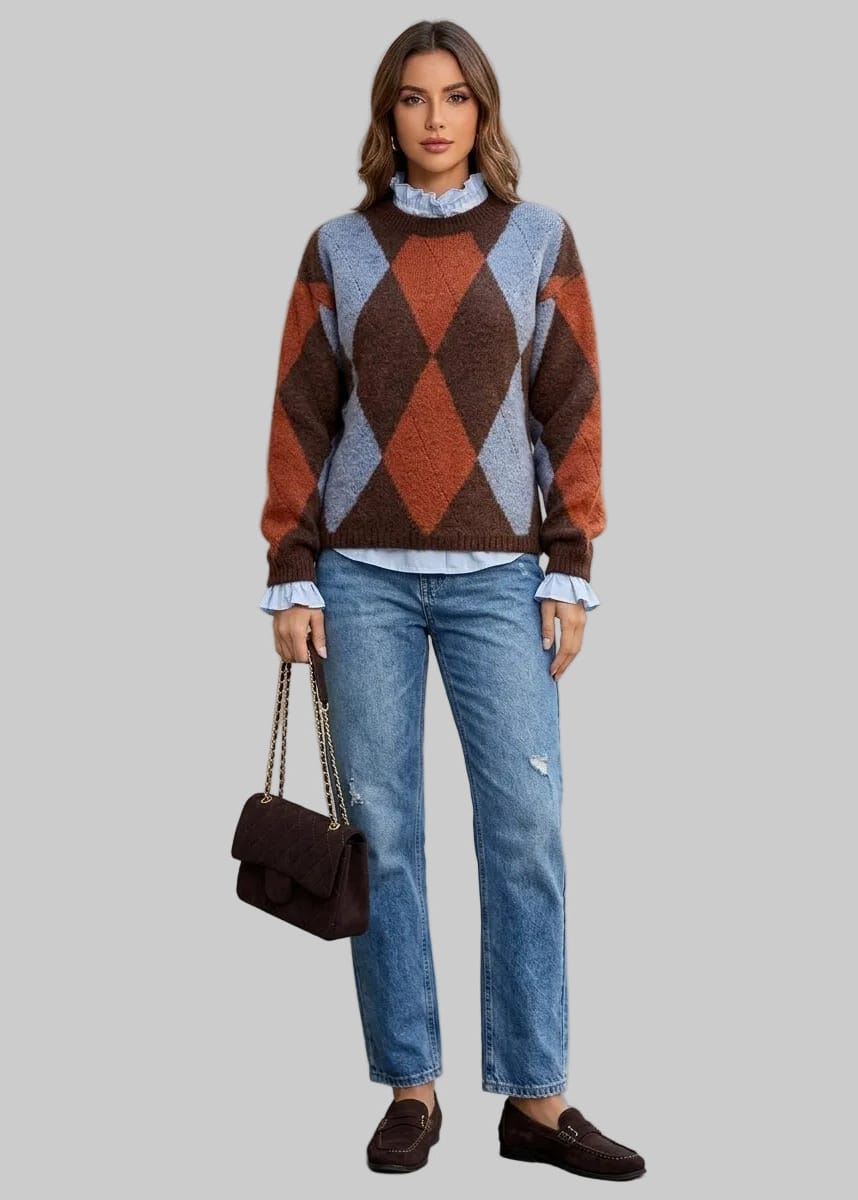 A woman stands outdoors against a light gray wall wearing a brown, rust, and light blue argyle sweater over a light blue ruffled blouse, paired with blue jeans and dark brown loafers. She is holding a dark brown handbag.