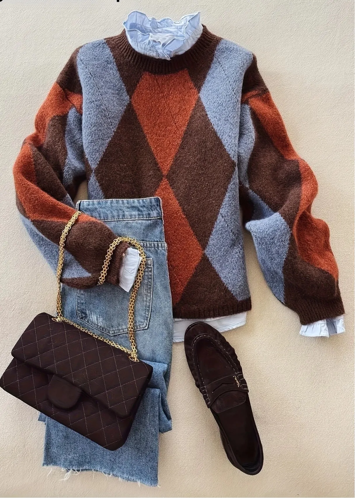 A styled flat-lay featuring a fuzzy argyle sweater in brown, rust orange, and light blue-gray, layered over a light blue ruffled blouse. It is paired with light-wash jeans, a dark brown quilted handbag with a gold chain, and a dark brown loafer, all arranged on a beige textured background.