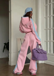 Person wearing a pink tracksuit with a light blue hood and holding a purple bag, standing in a room.