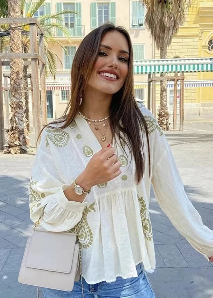 Woman in a white blouse with gold embroidery standing outdoors with palm trees and buildings in the background.