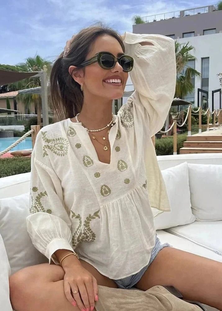 Woman wearing a white embroidered blouse and denim shorts sitting on a white couch with a pool and palm trees in the background.