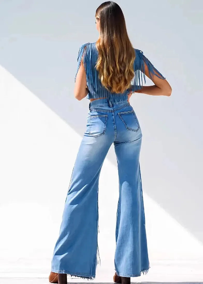 A woman shown from behind wearing a light blue denim jumpsuit with fringe details on the sleeves and back, paired with brown wedge sandals, set against a bright white background with diagonal shadows.