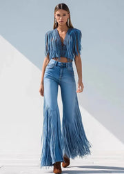A woman models a two-piece blue denim set featuring a fringed short-sleeve crop top and matching fringed flare jeans, styled with brown boots against a minimalist white and light blue background.