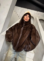 A person wearing an oversized dark brown fur-like zip jacket, light blue jeans, and a beige cap, standing on concrete stairs in a minimalist industrial interior.