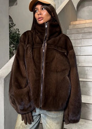 A woman wearing a dark brown mink jacket with a high collar and front zipper, paired with light blue jeans, brown gloves, and a beige cap, standing in front of a gray stone staircase.