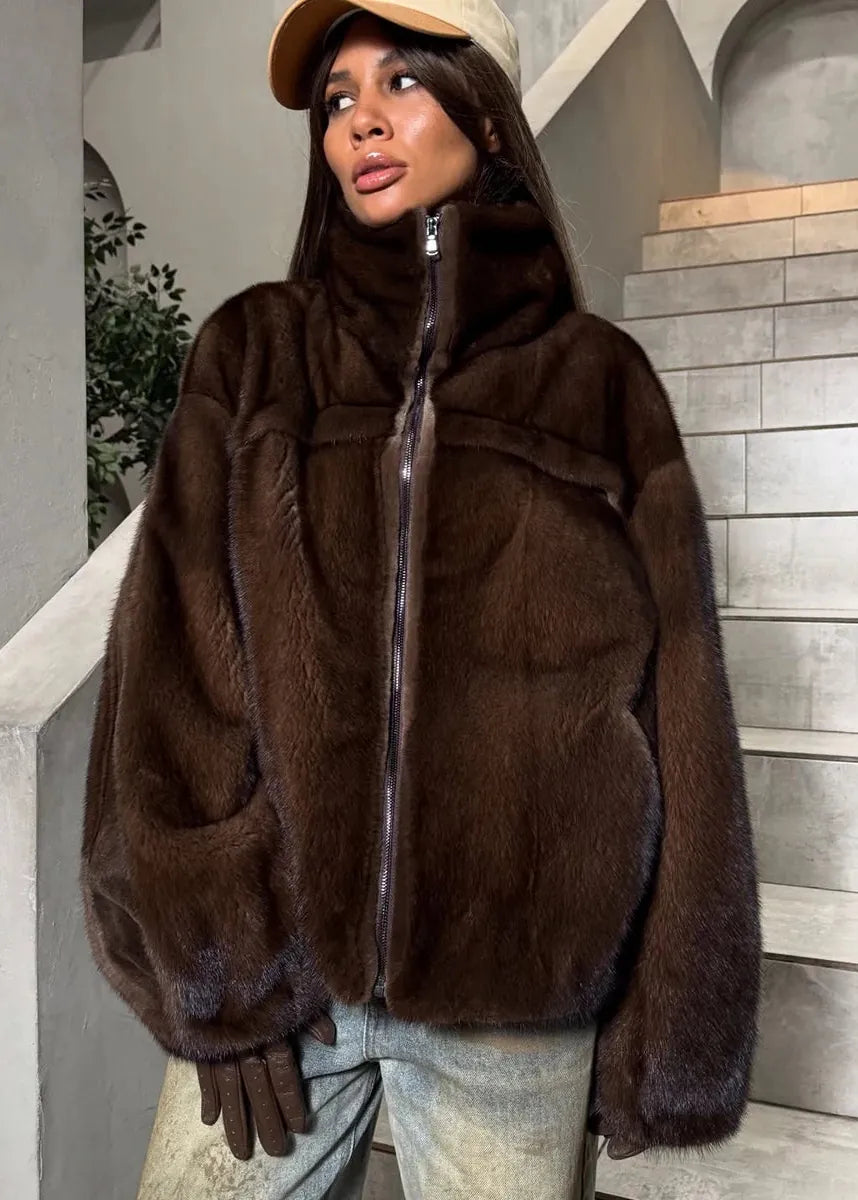 A woman wearing a dark brown mink jacket with a high collar and front zipper, paired with light blue jeans, brown gloves, and a beige cap, standing in front of a gray stone staircase.