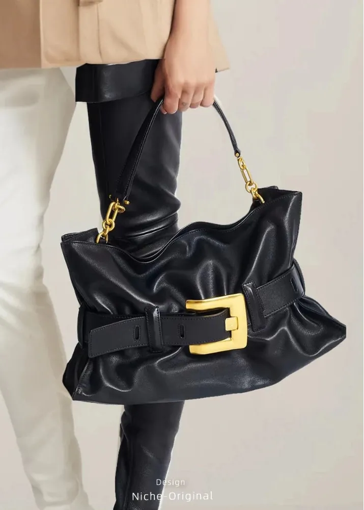 A black leather slouchy shoulder bag with a wide strap and a prominent gold buckle, held by a model wearing beige pants and black boots.