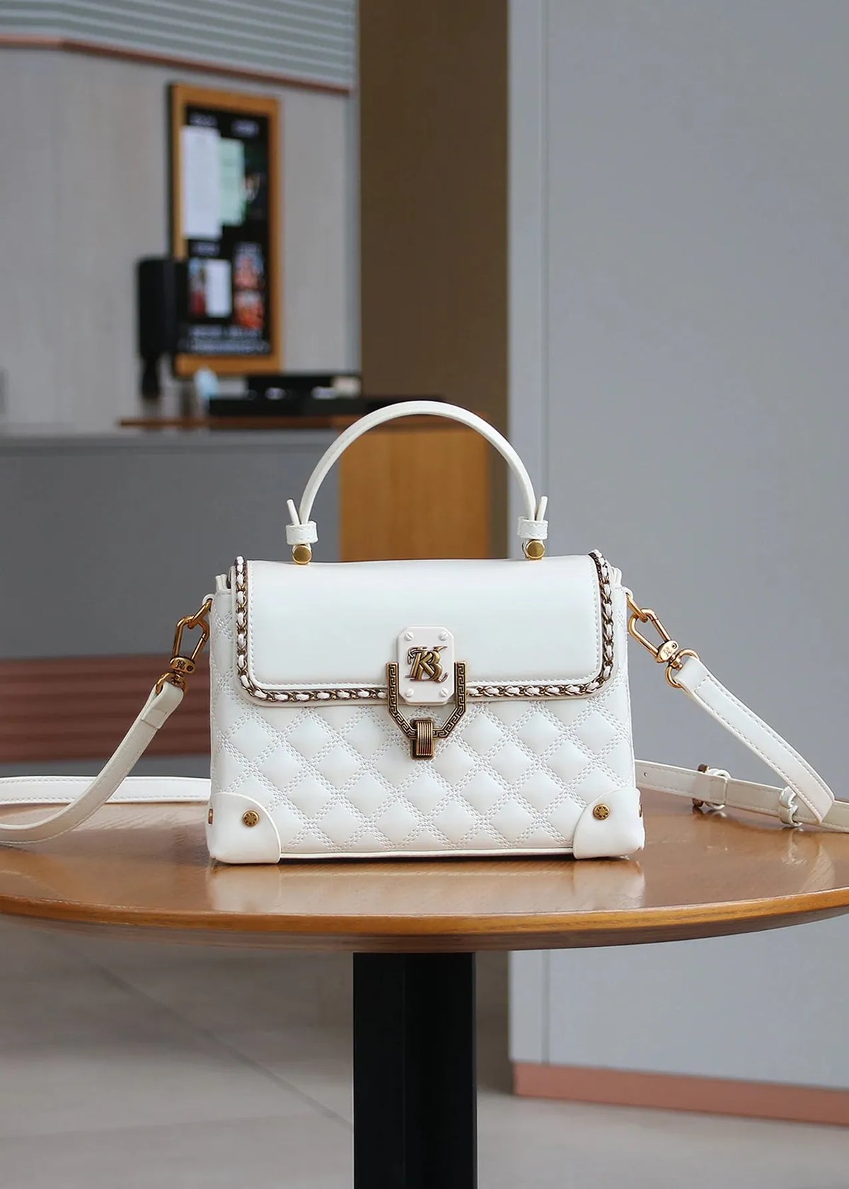 A white quilted leather handbag with gold-tone hardware and a top handle, displayed on a round wooden table in an indoor setting.