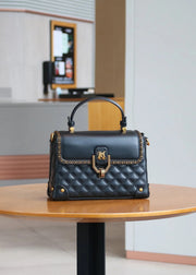 A black quilted leather handbag with gold-tone hardware and a top handle, displayed on a round wooden table in a modern indoor setting.