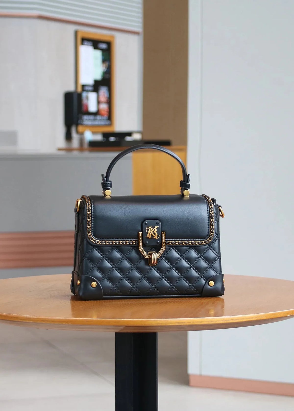 A black quilted leather handbag with gold-tone hardware and a top handle, displayed on a round wooden table in a modern indoor setting.