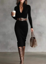 Woman in a black dress with brown belt and boots, holding a coffee cup and a clear handbag against a gray background.