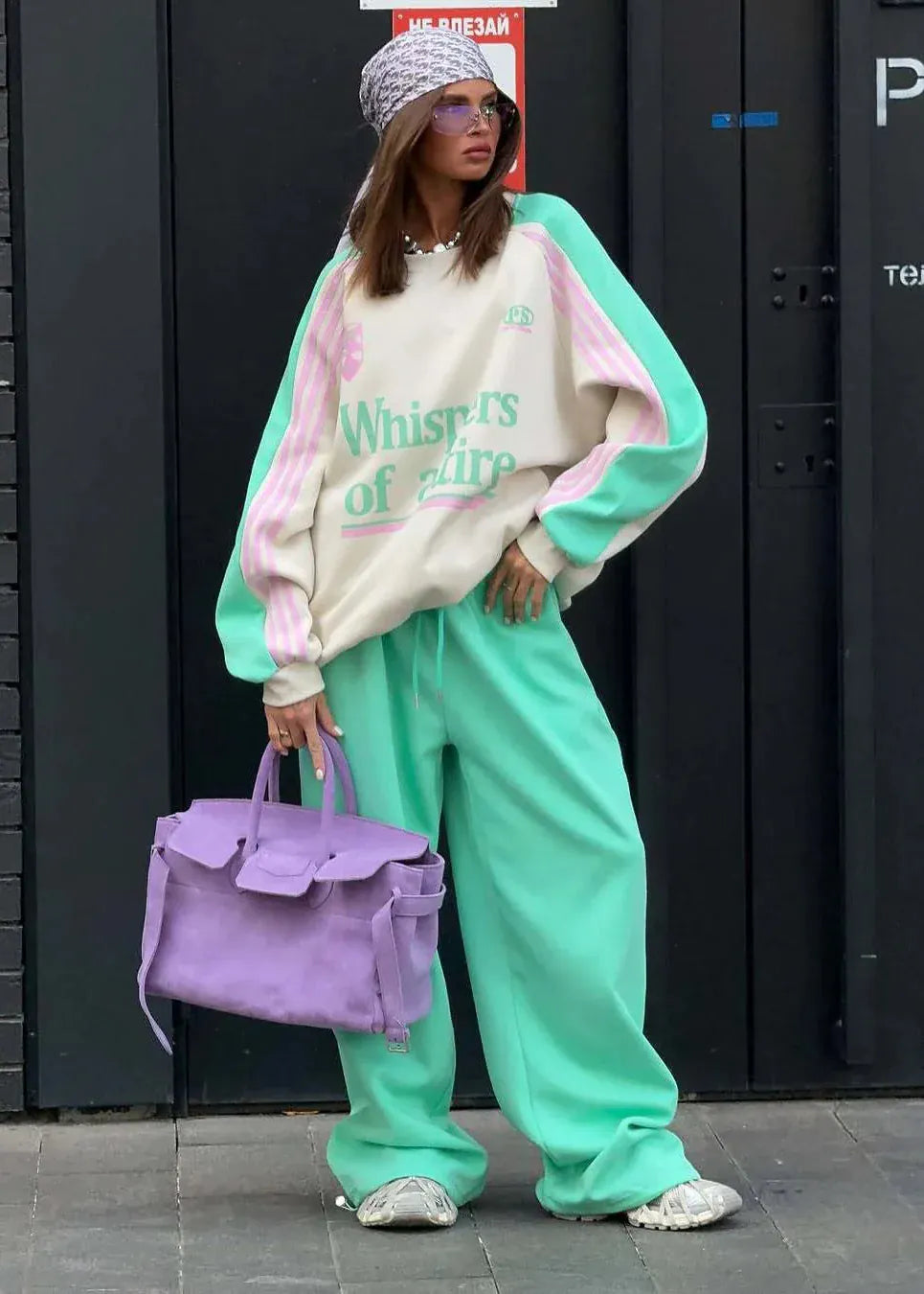 Person wearing a colorful outfit with a purple bag against a black wall.