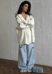 A woman stands indoors wearing an oversized cream knit cardigan with small red and white embroidered details, light-wash distressed jeans, and a small blue-gray shoulder bag. The background is a textured off-white wall with a wood floor.