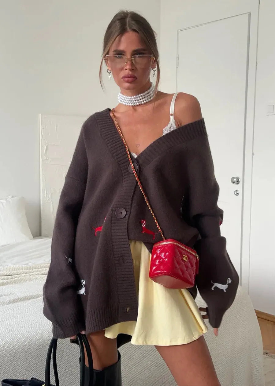 Woman wearing a dark brown cardigan over a white top and yellow skirt, holding a red handbag in a bedroom.