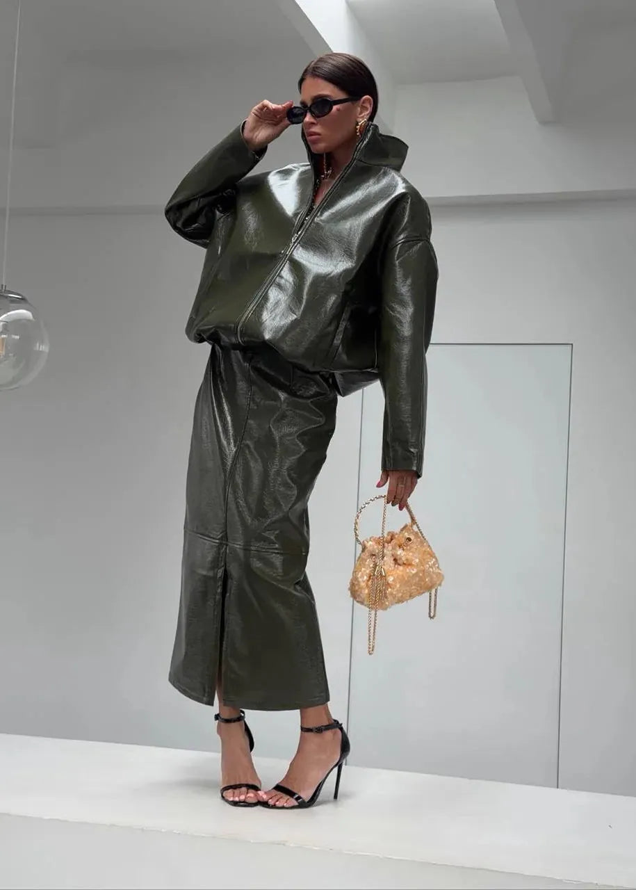 A model stands on a white platform in a minimalist studio setting, wearing a matching olive green faux-leather jacket and skirt set, black strappy heels, and holding a small beige patterned handbag.