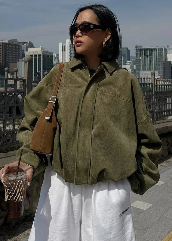 Woman in a green jacket and white pants holding a drink with a cityscape in the background