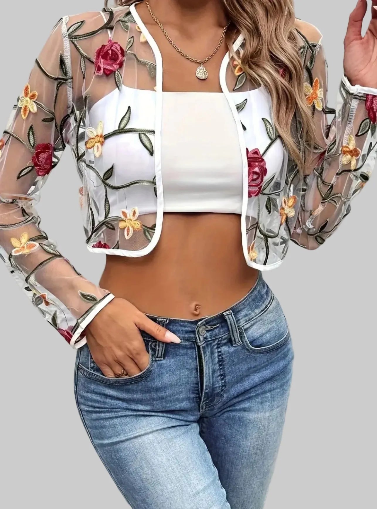 A woman wearing a sheer floral embroidered jacket over a white crop top and blue jeans, standing against a textured stone wall.