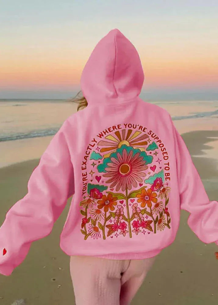 Person wearing a pink hoodie with floral design and text on a beach at sunset